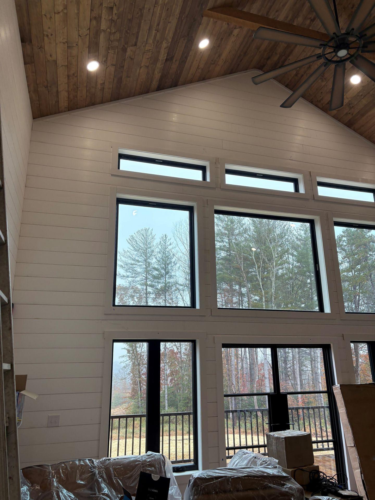 Interior view of a room with large windows, a high wooden ceiling, and trees visible outside.