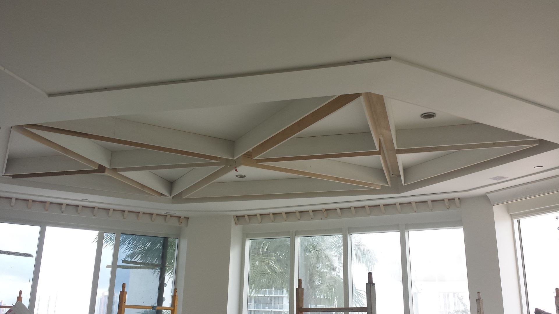 Interior view of a room with a geometric ceiling design under construction and large windows overlooking trees.